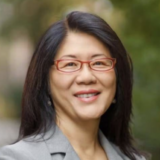 A person with long dark hair, red glasses, and a gray blazer poses outdoors for a photo